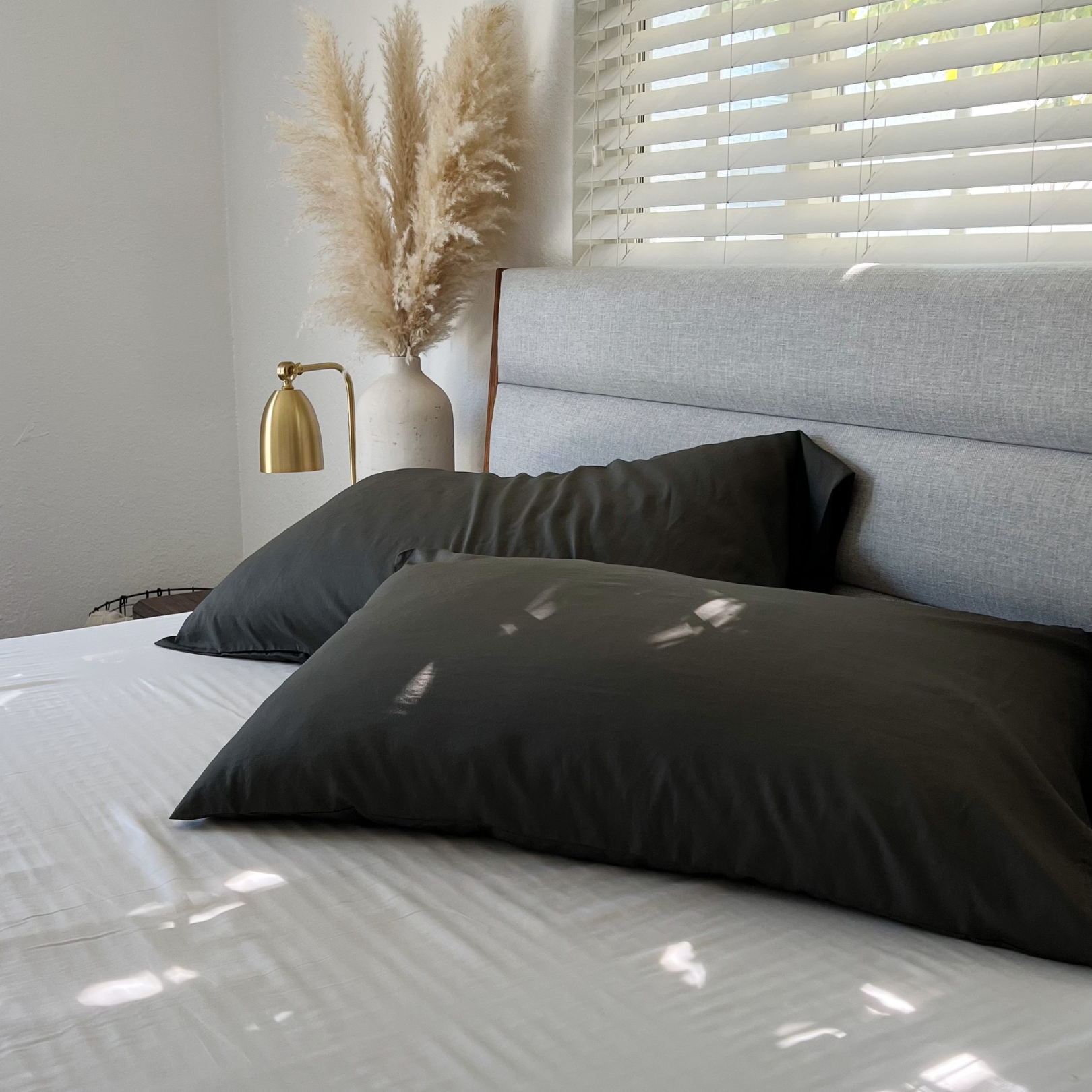 Dark grey Copper Infused Bamboo Pillowcase set on a white bed, with pampas grass and a gold lamp.